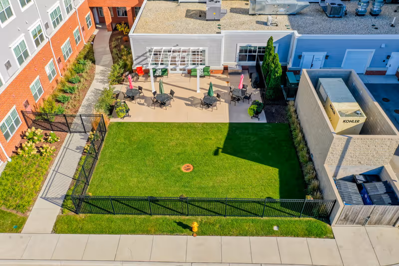 Aerial view of a fenced grassy courtyard and patio with tables, umbrellas, and landscaping behind a brick-and-siding senior living building.