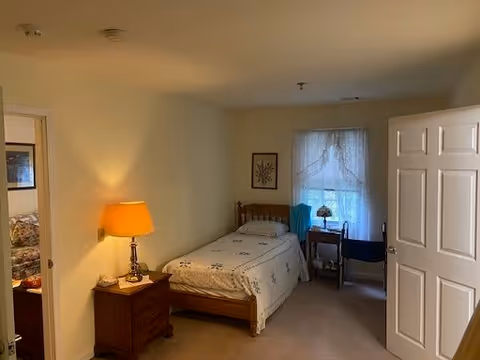 A simple bedroom with a single wooden bed covered with a white bedspread featuring a floral pattern. Next to the bed is a wooden nightstand with a lamp emitting warm light. There is a window with sheer curtains letting in natural light, and a small desk with a chair and a lamp is positioned in front of the window. The walls are light-colored, and a framed picture hangs on the wall above the bed. The door to the room is open, showing a glimpse of another room with a floral couch.