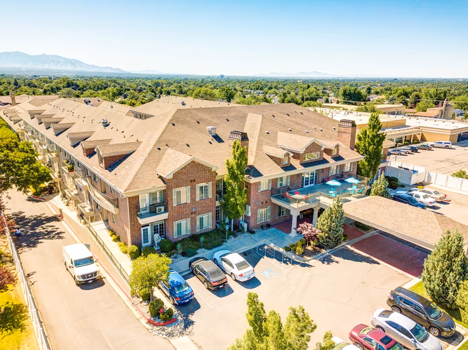 Aerial view of a large brick senior living facility named Escalante at Coventry, surrounded by trees and parking spaces with several cars. The building has multiple floors with balconies and a covered entrance area. Mountains and a clear blue sky are visible in the background.