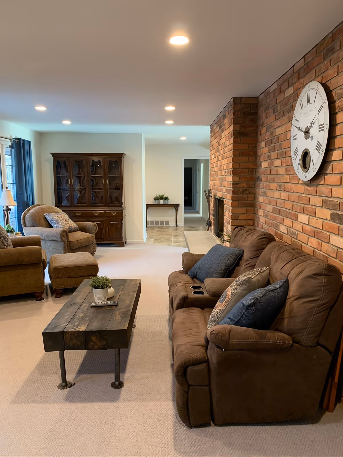 Cozy living room with a brown reclining sofa and pillows, wooden coffee table, armchairs, a brick fireplace wall with a large clock, and a display cabinet in the background.