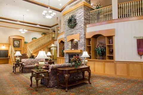 A spacious and warmly decorated senior living facility common area with floral upholstered sofas, wooden tables with lamps, a large stone fireplace adorned with a wreath, wooden staircase with railings, and built-in shelves with decorative plants and flowers.