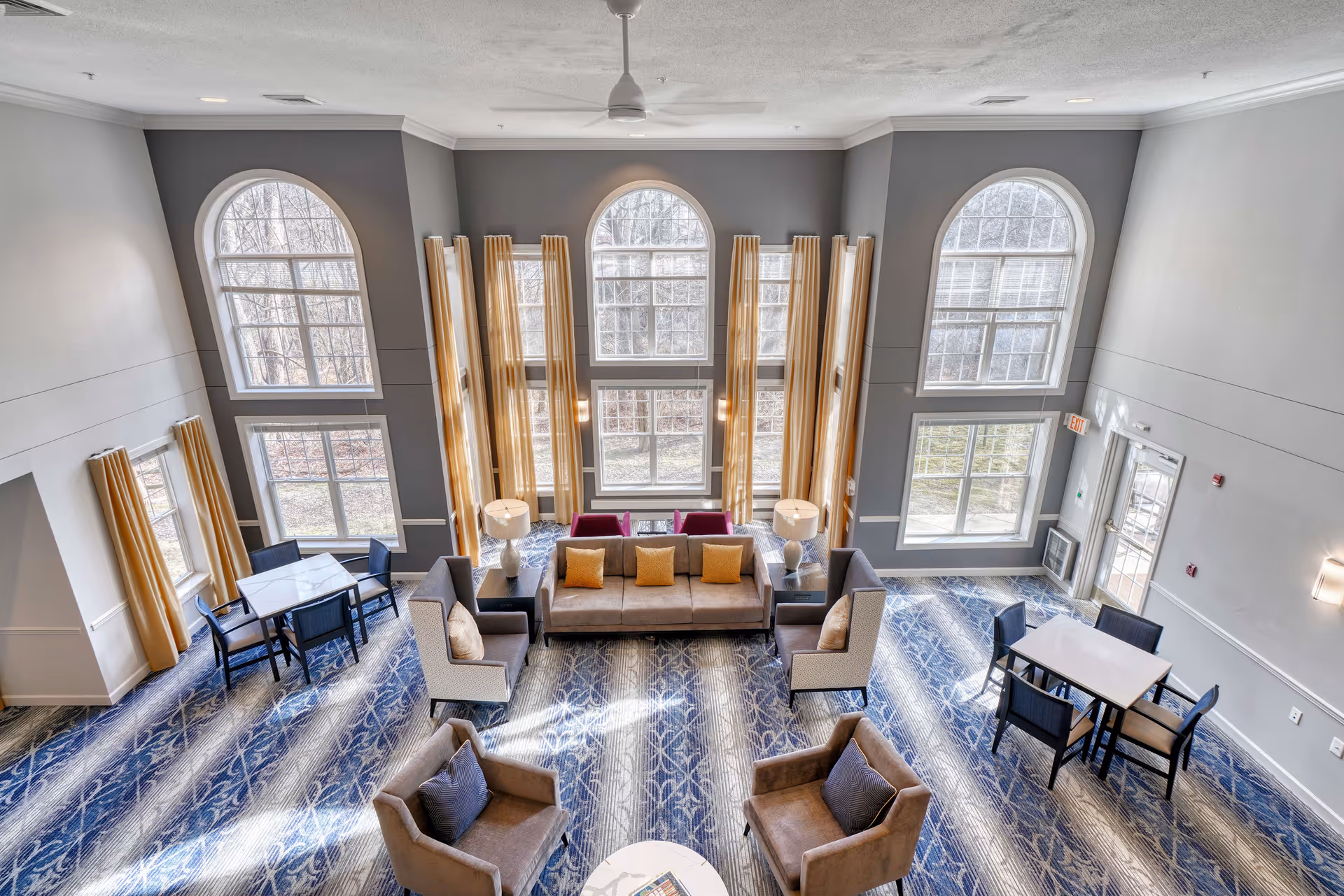 Two-story senior living lounge with large arched windows, sofas and armchairs arranged around tables and blue patterned carpet.