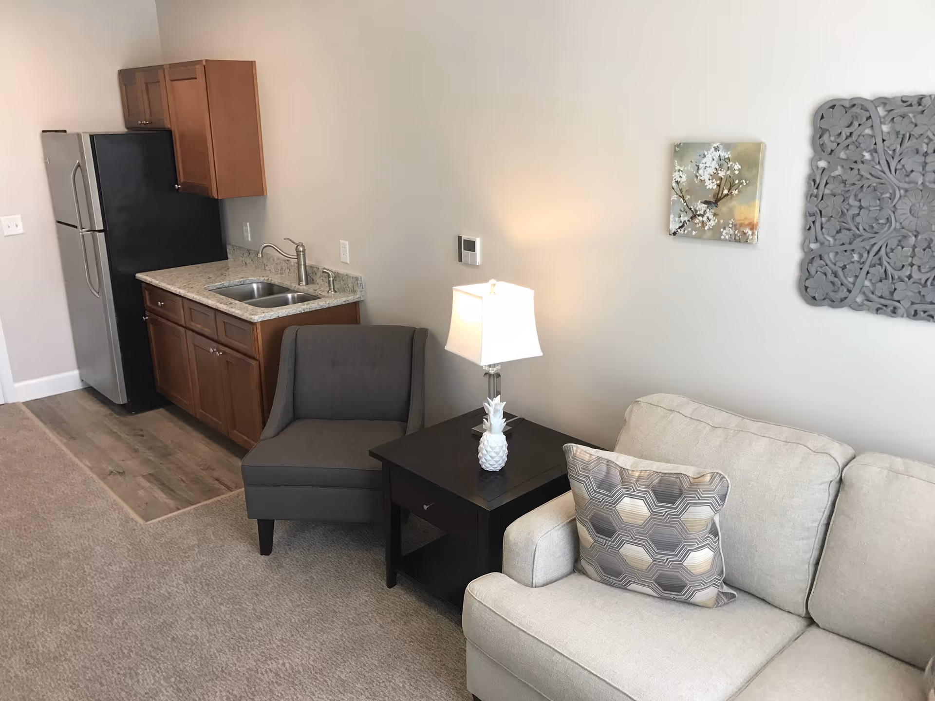 A small living area with a beige couch featuring a patterned pillow, a dark wooden side table with a white pineapple-shaped lamp, a gray upholstered chair, and a kitchenette with wooden cabinets, a stainless steel refrigerator, and a sink. The walls are light-colored with two decorative wall hangings.