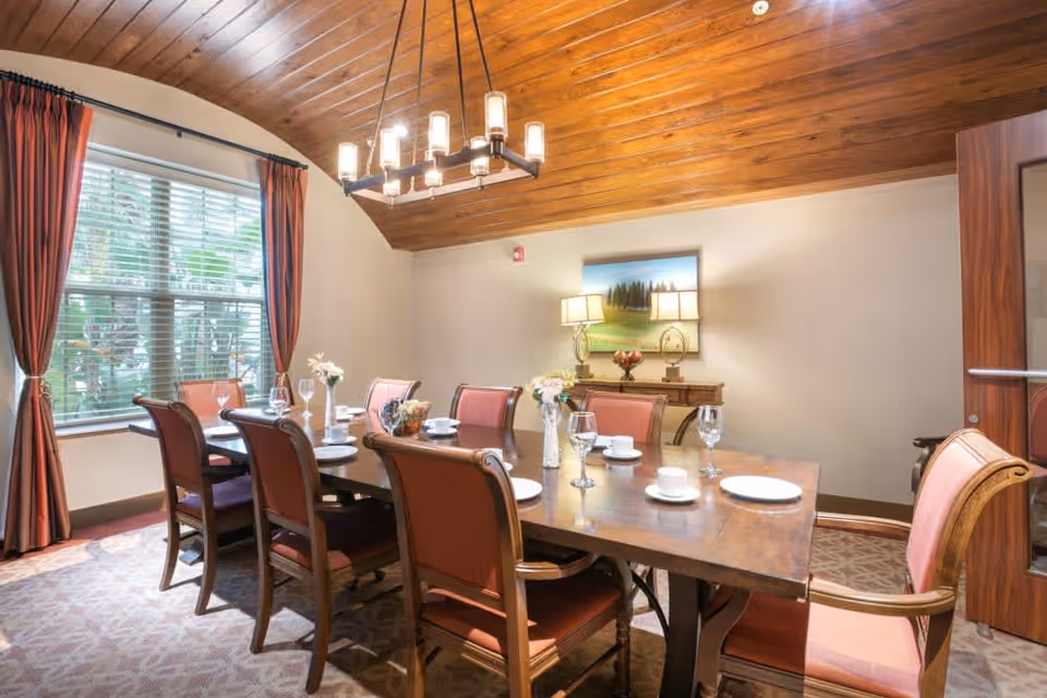 A well-lit dining room with a wooden table set for eight people. The table has plates, cups, wine glasses, and small flower vases. The room features a wooden ceiling, a large window with red curtains, a sideboard with two lamps and a painting above it, and a carpeted floor.
