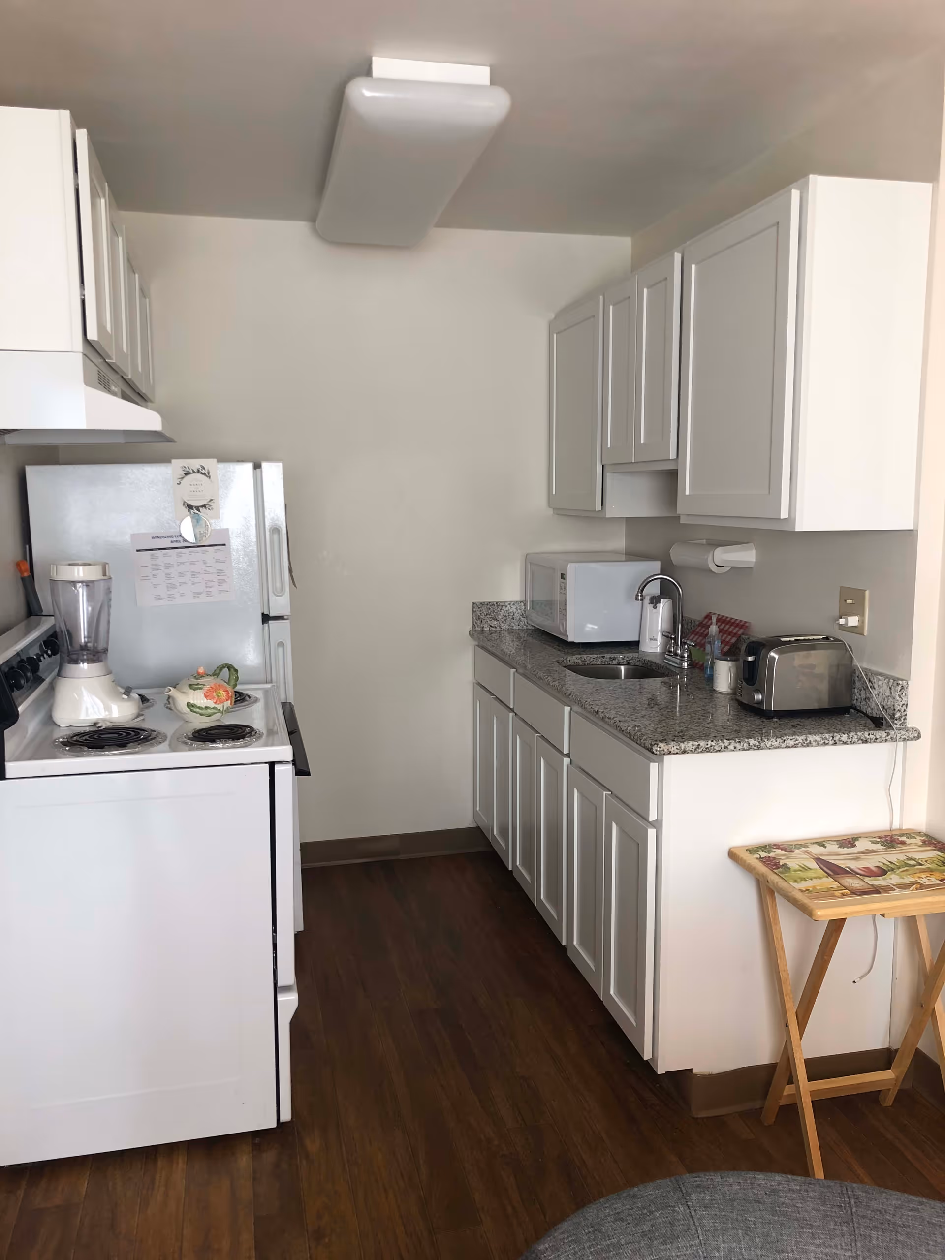 A small kitchen with white cabinets, a granite countertop, a white stove with a blender and a decorative teapot on it, a white refrigerator with papers attached, a microwave, a toaster, a sink with a faucet, and a small wooden folding table with a decorative top. The floor is wooden and the walls are plain white.