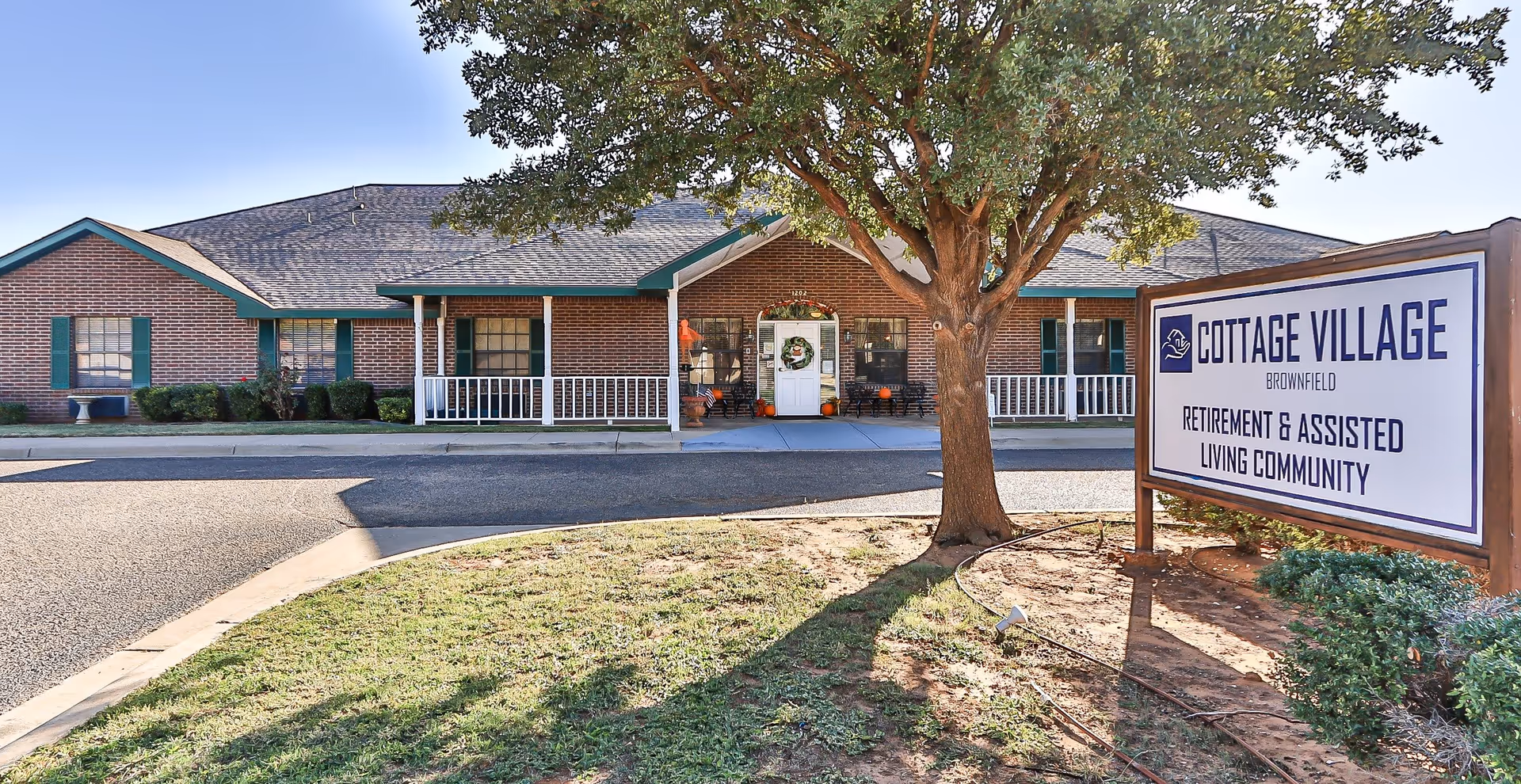 Front exterior of Cottage Village of Brownfield assisted living building with a tree, porch entrance, and a large sign.