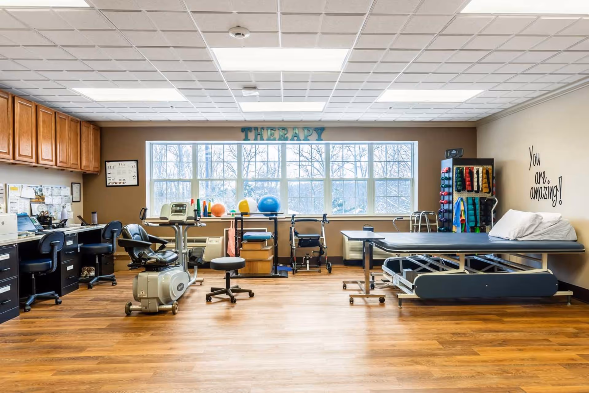 A bright therapy room with large windows letting in natural light. The room features exercise equipment including a stationary bike, therapy balls, weights, and a treatment table with pillows. There are desks and chairs along the left wall with various papers and a computer. The wall above the windows has the word 'THERAPY' displayed, and on the right wall, there is a motivational quote saying 'You are amazing!'.