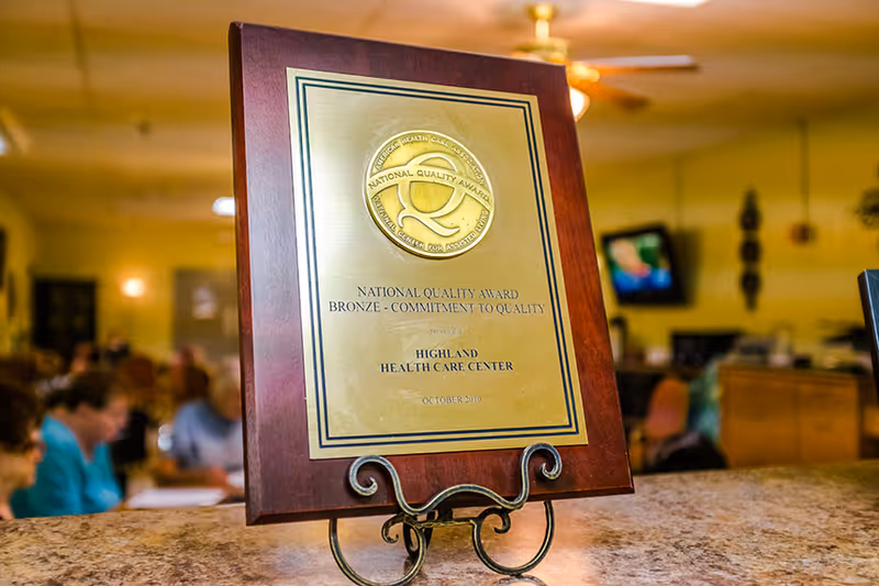 A close-up of a National Quality Award plaque for Highland Health Care Center, displayed on a stand on a countertop with a blurred background showing people and a television in a common area.