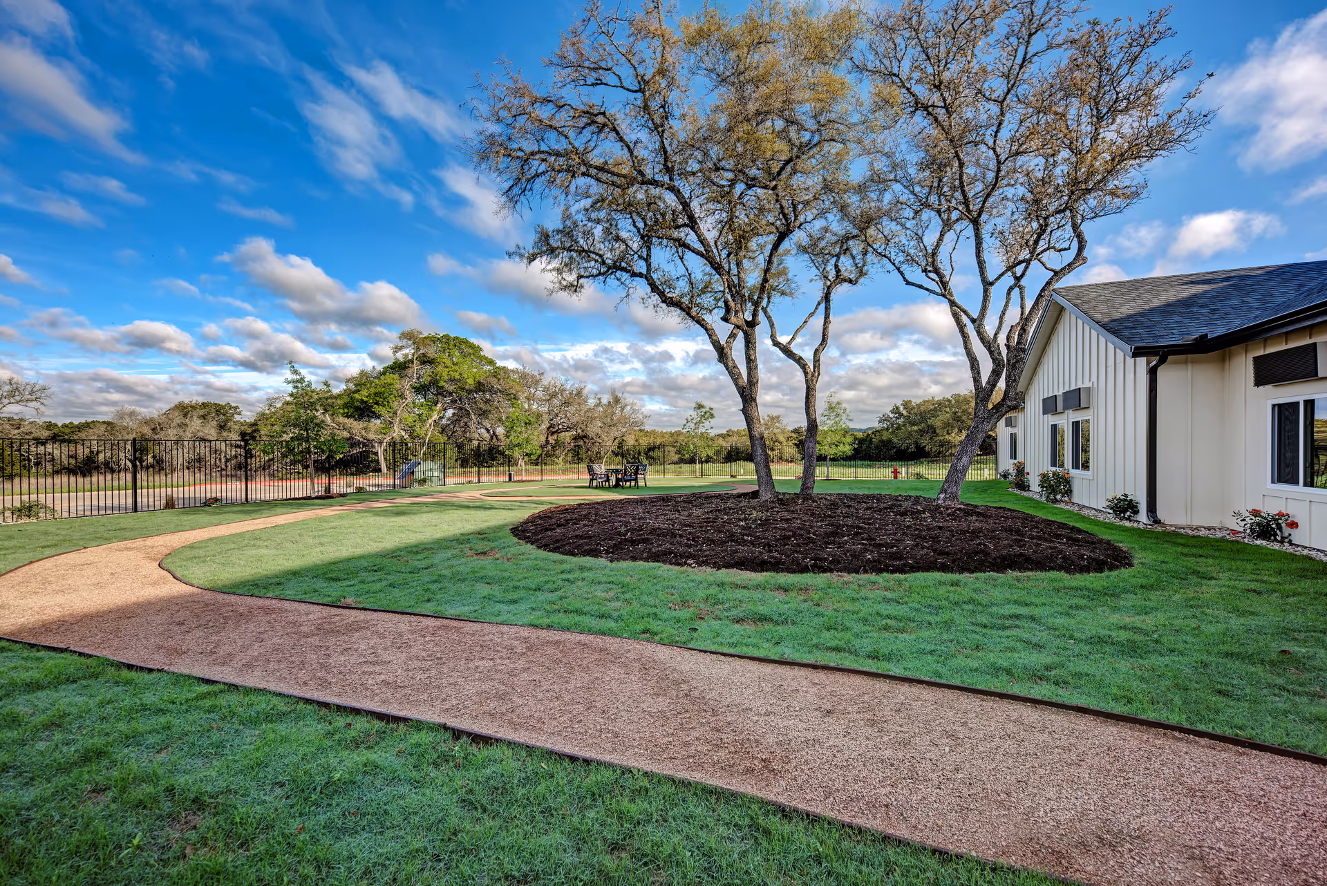 A landscaped outdoor area at Symphony of Wimberley featuring a curved walking path, green grass, several trees with bare branches, and a building with white siding and black roof on the right side. The sky is blue with scattered clouds.