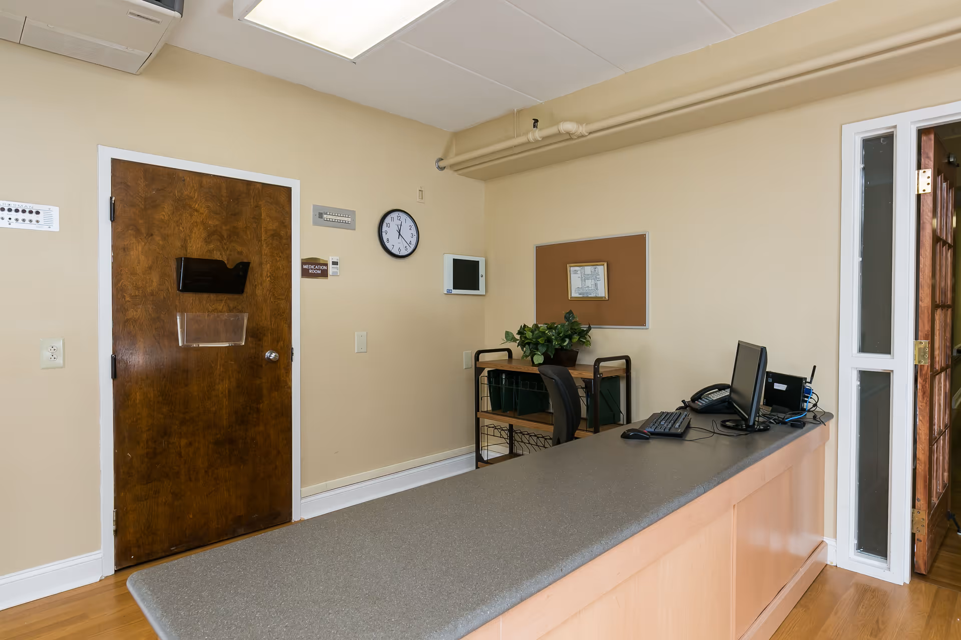 Reception desk area inside a facility with a computer, keyboard, mouse, and telephone on the counter. Behind the desk is a wooden door labeled 'Medication Room,' a wall clock, a small bulletin board, and a cart with a potted plant and some files. The walls are beige and the floor is wooden.