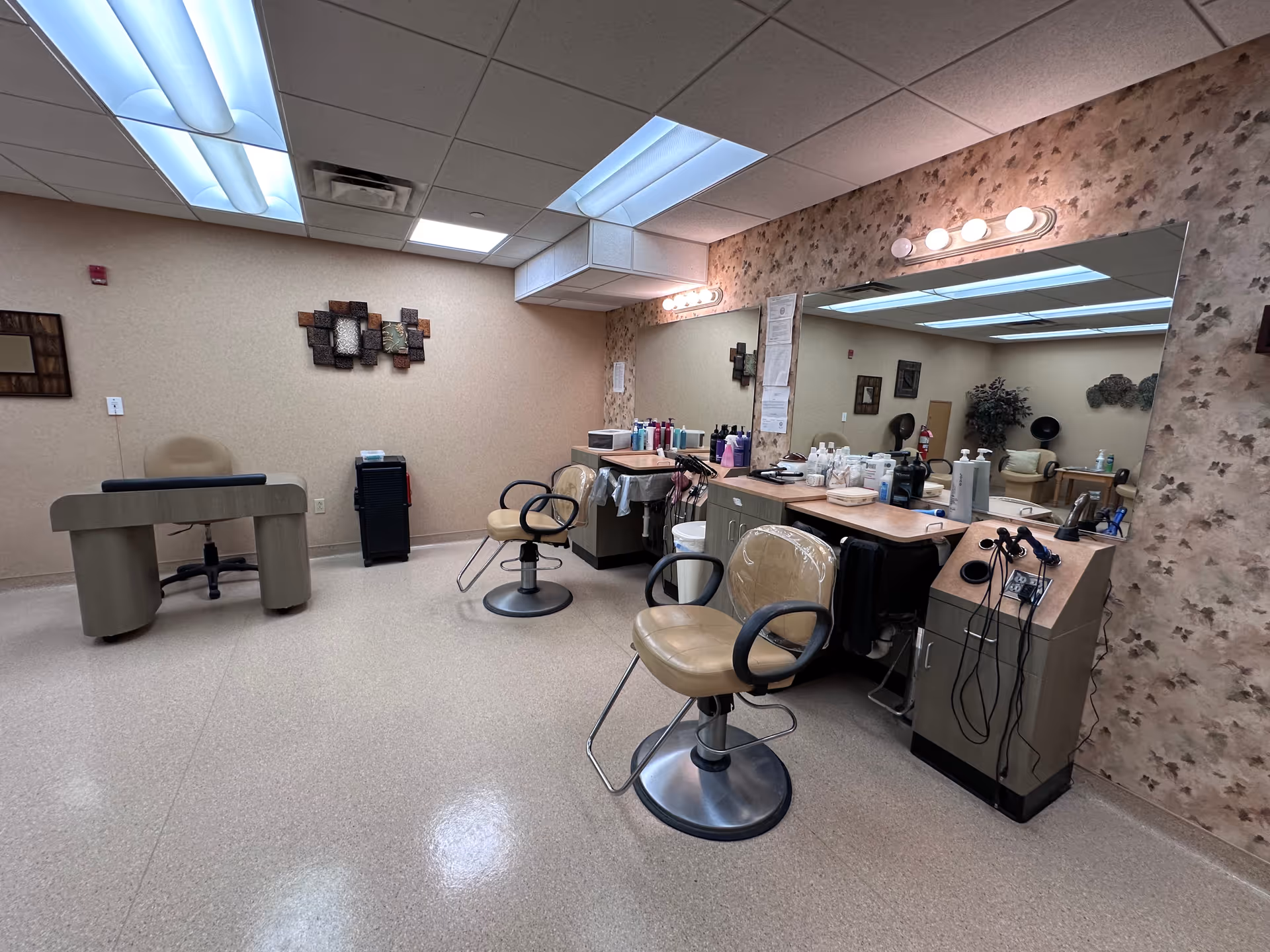 Interior view of a salon area in a senior living facility with two salon chairs in front of a large mirror, various hair care products on the counter, and a small desk with a chair in the background. The room has beige walls, a patterned accent wall, and fluorescent ceiling lights.