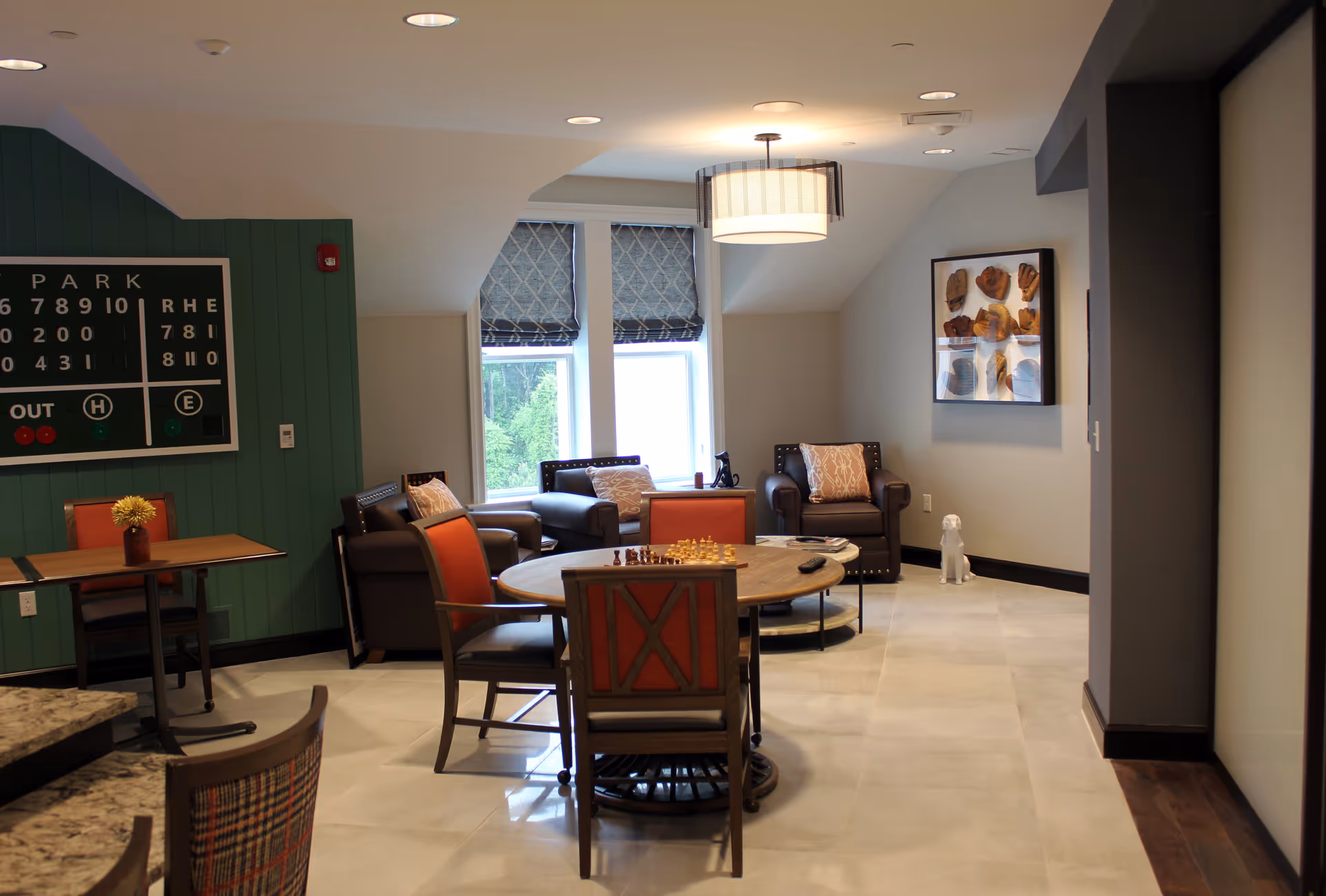 A cozy lounge area with a round wooden table in the center featuring a chess set. Surrounding the table are chairs with orange cushions. In the background, there are three brown leather armchairs with patterned pillows, two windows with patterned blinds, and a wall-mounted display of vintage baseball gloves. The walls are painted in neutral tones with one green accent wall that has a scoreboard mounted on it. The floor is tiled, and a modern ceiling light fixture hangs above the table.