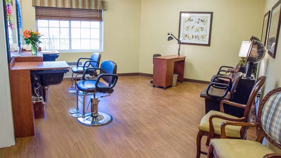 Bright salon-style room with barber chairs, styling stations, and waiting chairs near a large window.