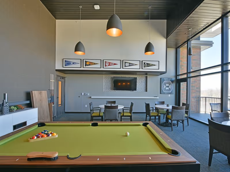 Bright communal lounge/game room with a pool table in the foreground, round tables and chairs, a wall-mounted TV, pendant lights, and large floor-to-ceiling windows.