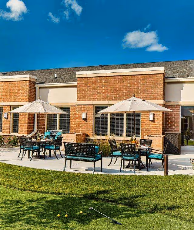 Outdoor patio with tables, chairs, umbrellas and a grassy lawn in front of a brick building.