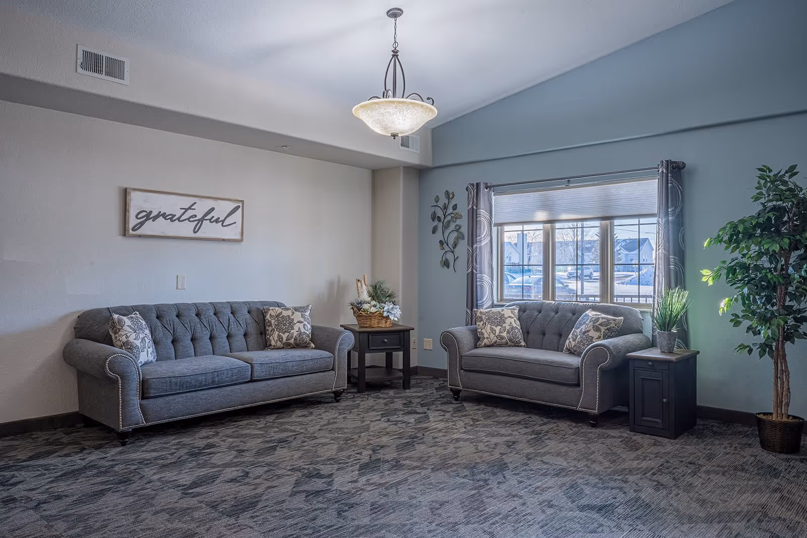 A cozy living room with two gray tufted sofas adorned with floral patterned pillows. Between the sofas is a small wooden side table with a decorative basket of plants. A large window with patterned curtains lets in natural light, and a potted plant stands to the right. A wall decoration with the word 'grateful' hangs above one sofa, and a ceiling light fixture illuminates the room.