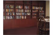 Interior view of a room with a large wooden bookshelf filled with books along the wall and a wooden dining table with chairs in the foreground.