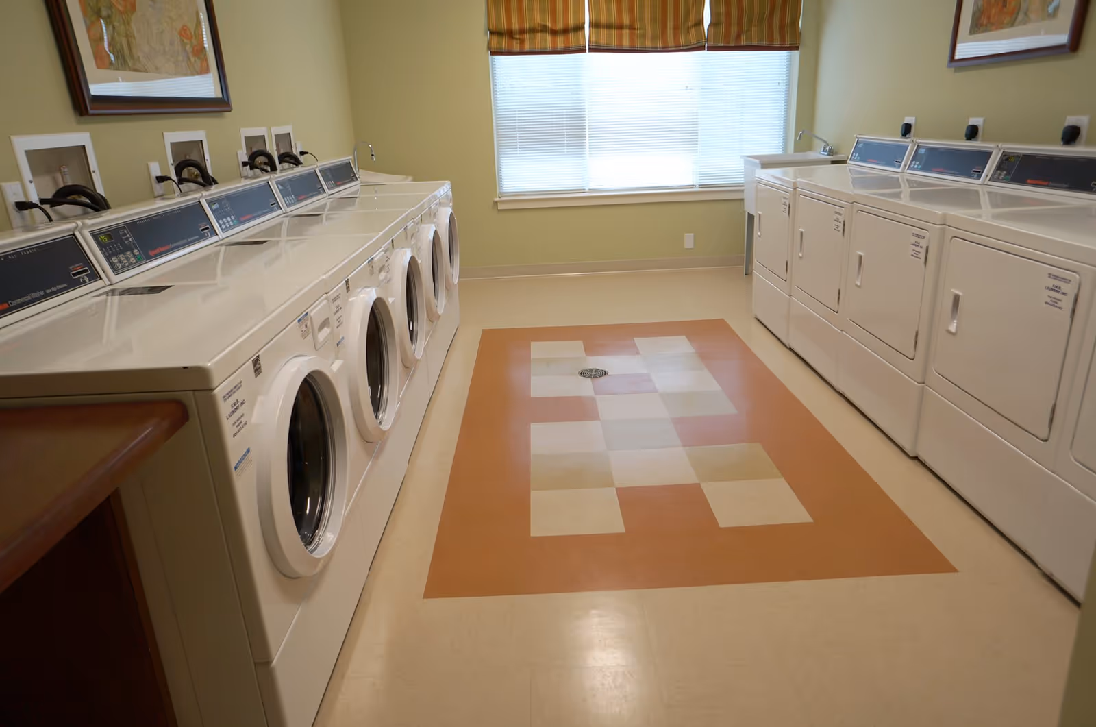 Laundry room with front-loading washers along the left, dryers on the right, and a patterned linoleum floor under a window at the far end.
