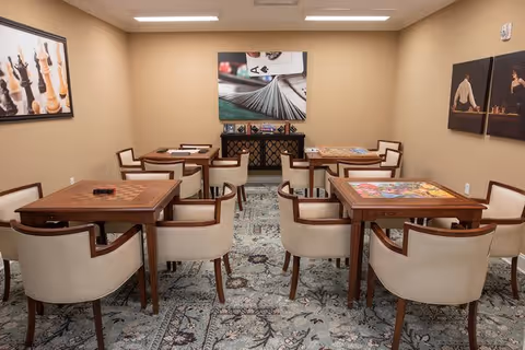 A well-lit game room with four wooden tables, each surrounded by four beige cushioned chairs. The tables have different board games and puzzles on them. The room has beige walls adorned with large framed pictures of chess pieces and playing cards. A patterned carpet covers the floor.