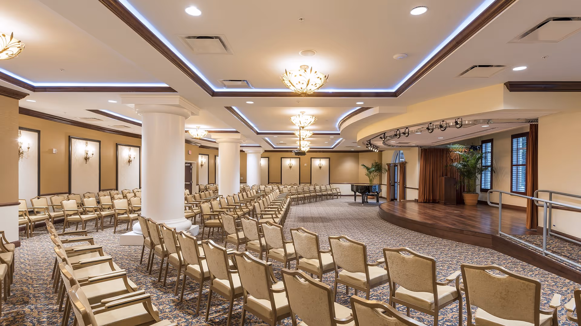 A large, elegant event room with rows of beige cushioned chairs arranged facing a wooden stage with a piano and potted plants. The room features decorative ceiling lights, white columns, and patterned carpet.