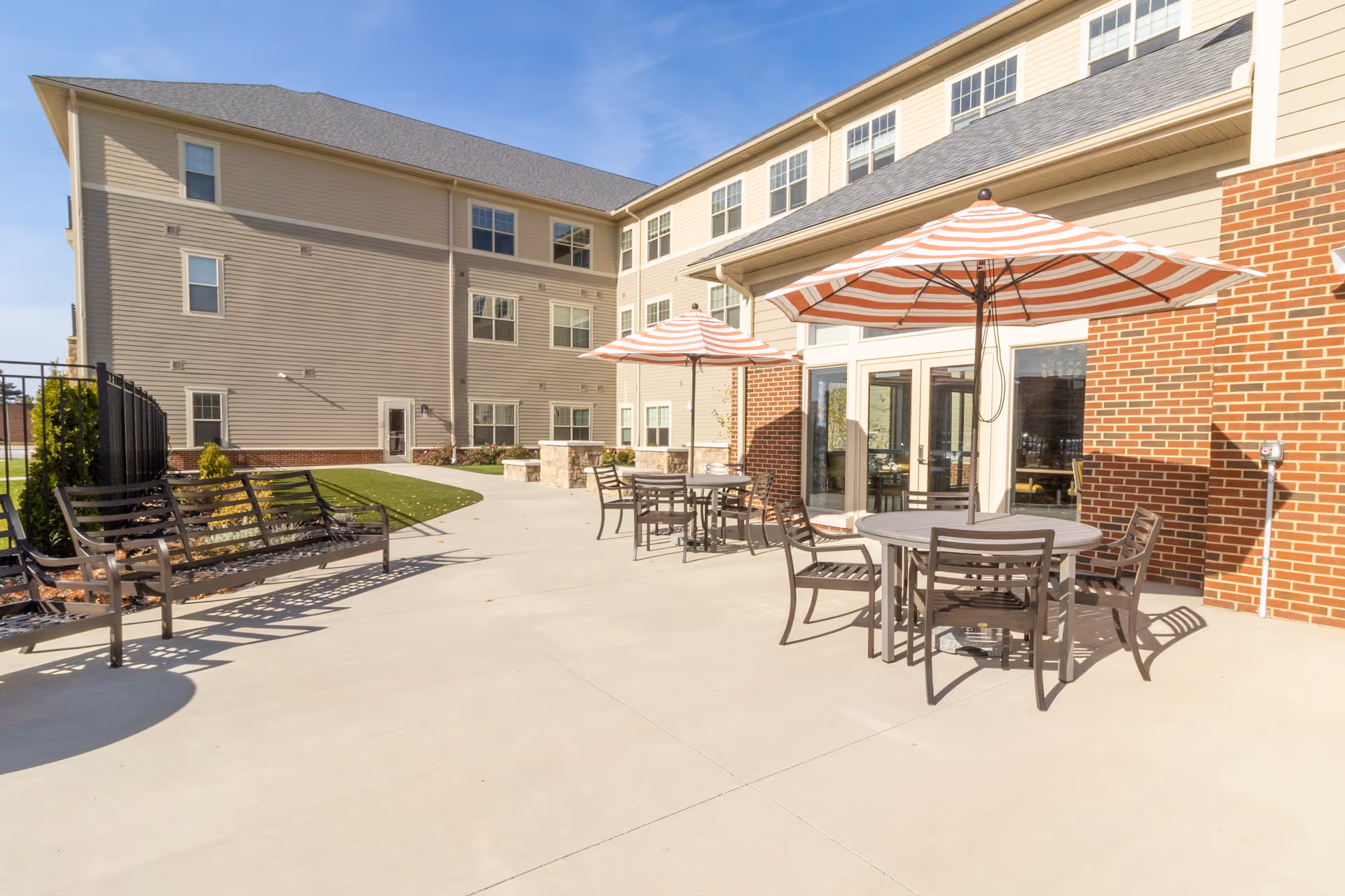 Outdoor patio with round tables, striped umbrellas, benches and a multi-story residential building in the background.