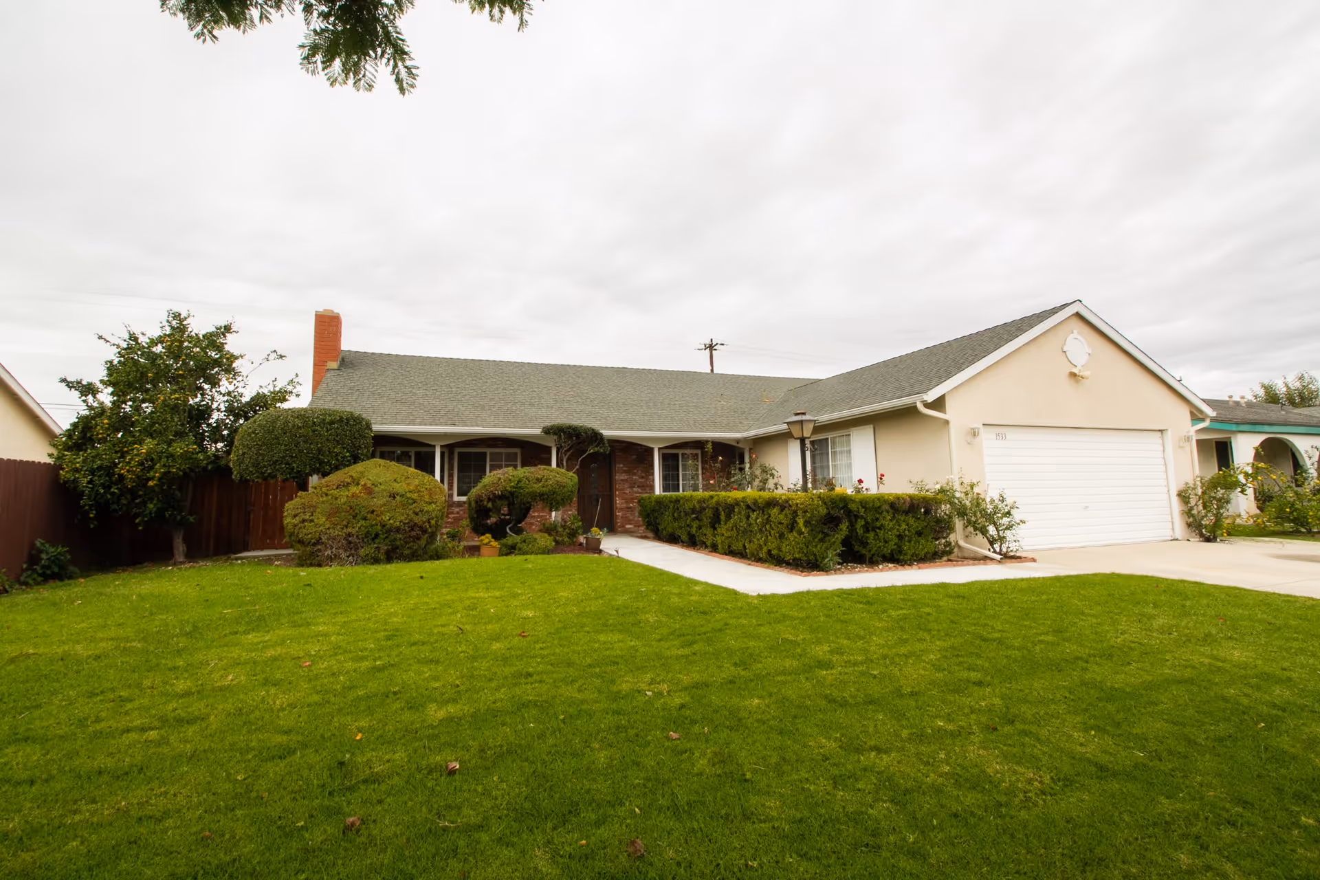 Single-story residential house with a green lawn, trimmed bushes, and a concrete driveway leading to a white garage door under an overcast sky.