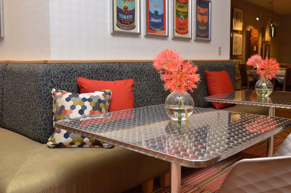 A cozy dining area with metal tables and cushioned bench seating adorned with colorful pillows. Each table has a small round glass vase with vibrant pink flowers. The wall behind the bench features framed artwork of Campbell's tomato soup cans in various colors.