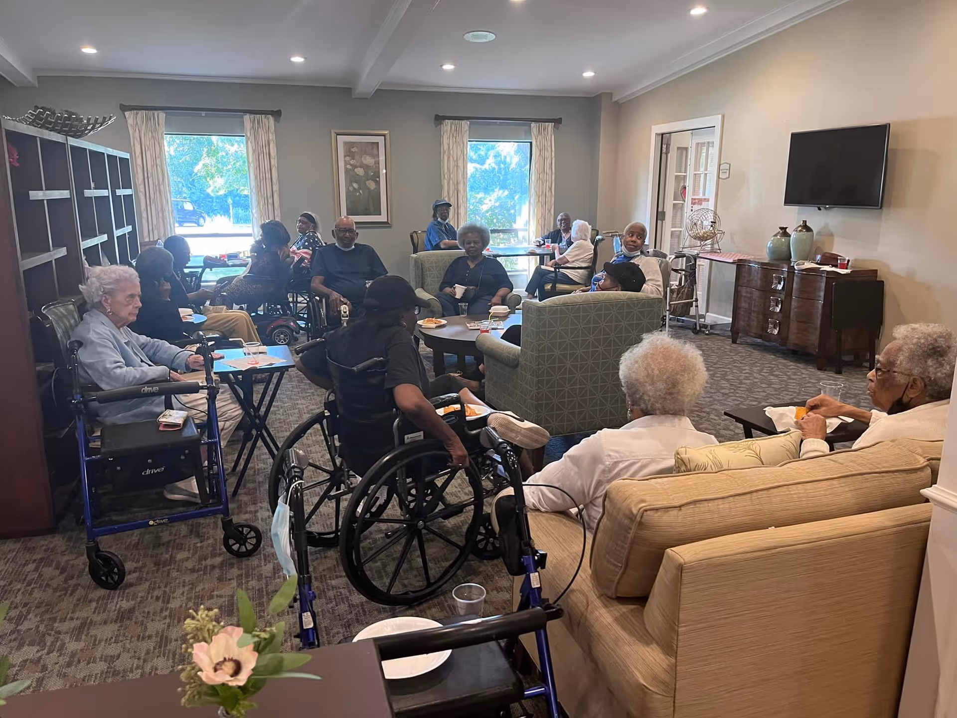 A group of elderly residents sitting and socializing in a well-lit common room with large windows. Some are seated in wheelchairs and others on armchairs and sofas arranged around a coffee table. The room has carpeted floors, a TV mounted on the wall, and decorative items on a sideboard.