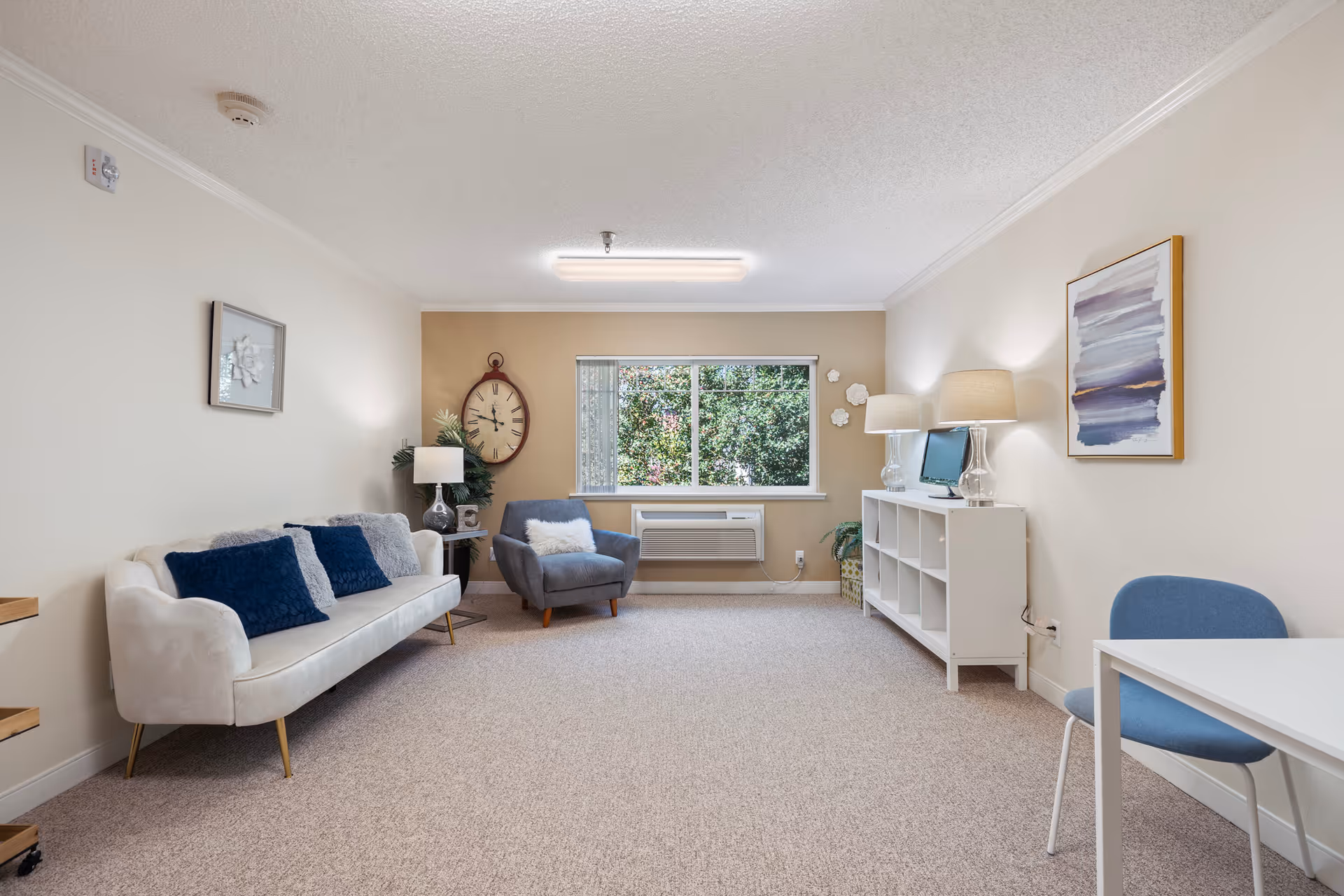Bright furnished living room with a white sofa and blue pillows, an armchair, window, and white shelving console with lamps.