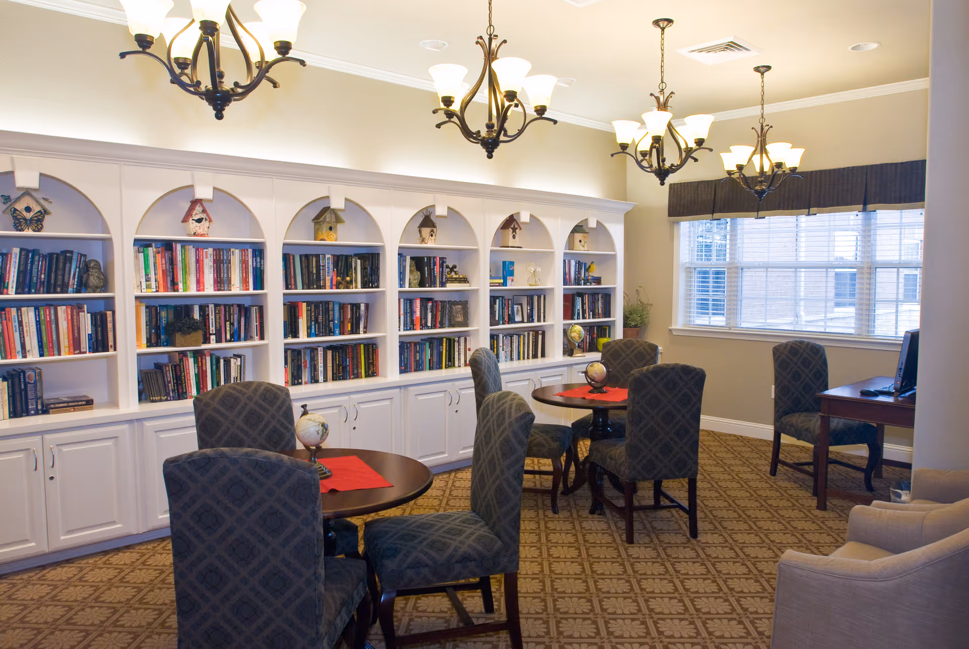 A cozy library or reading room with white built-in bookshelves filled with books and decorative birdhouses. There are round wooden tables with patterned upholstered chairs around them. The room is well-lit with multiple chandeliers and natural light coming through a large window with blinds. A desk with a computer is visible near the window.