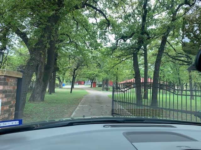 View from inside a vehicle looking through the windshield at a driveway lined with trees and a black metal gate that is partially open. There is a brick pillar on the left and green grass on both sides of the driveway, with red buildings visible in the background.