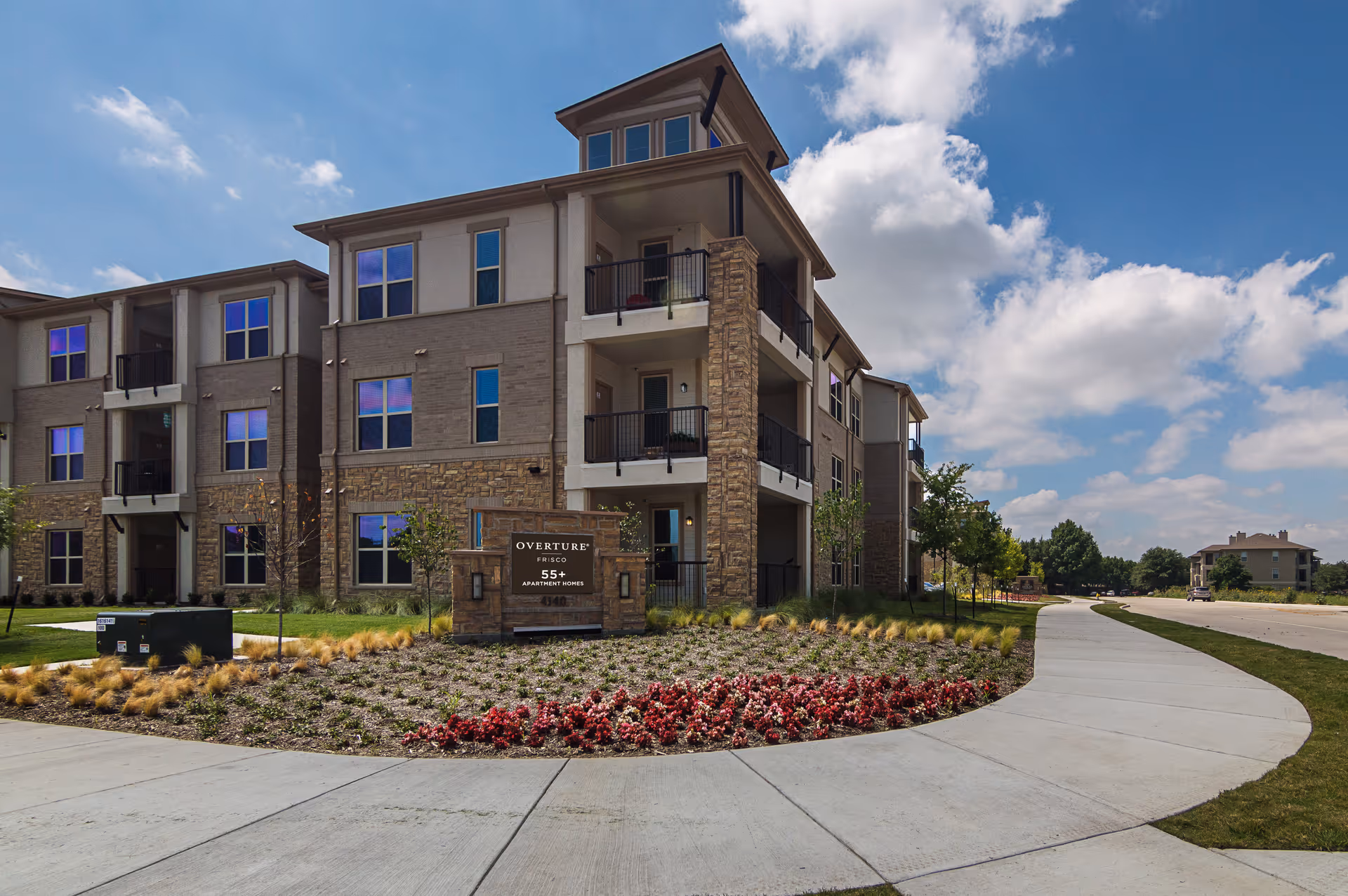 Exterior view of a modern three-story apartment building with balconies, a landscaped garden with red flowers and ornamental grasses, a curved sidewalk, and a sign that reads 'Overture Frisco 55+ Apartment Homes'. The sky is partly cloudy.