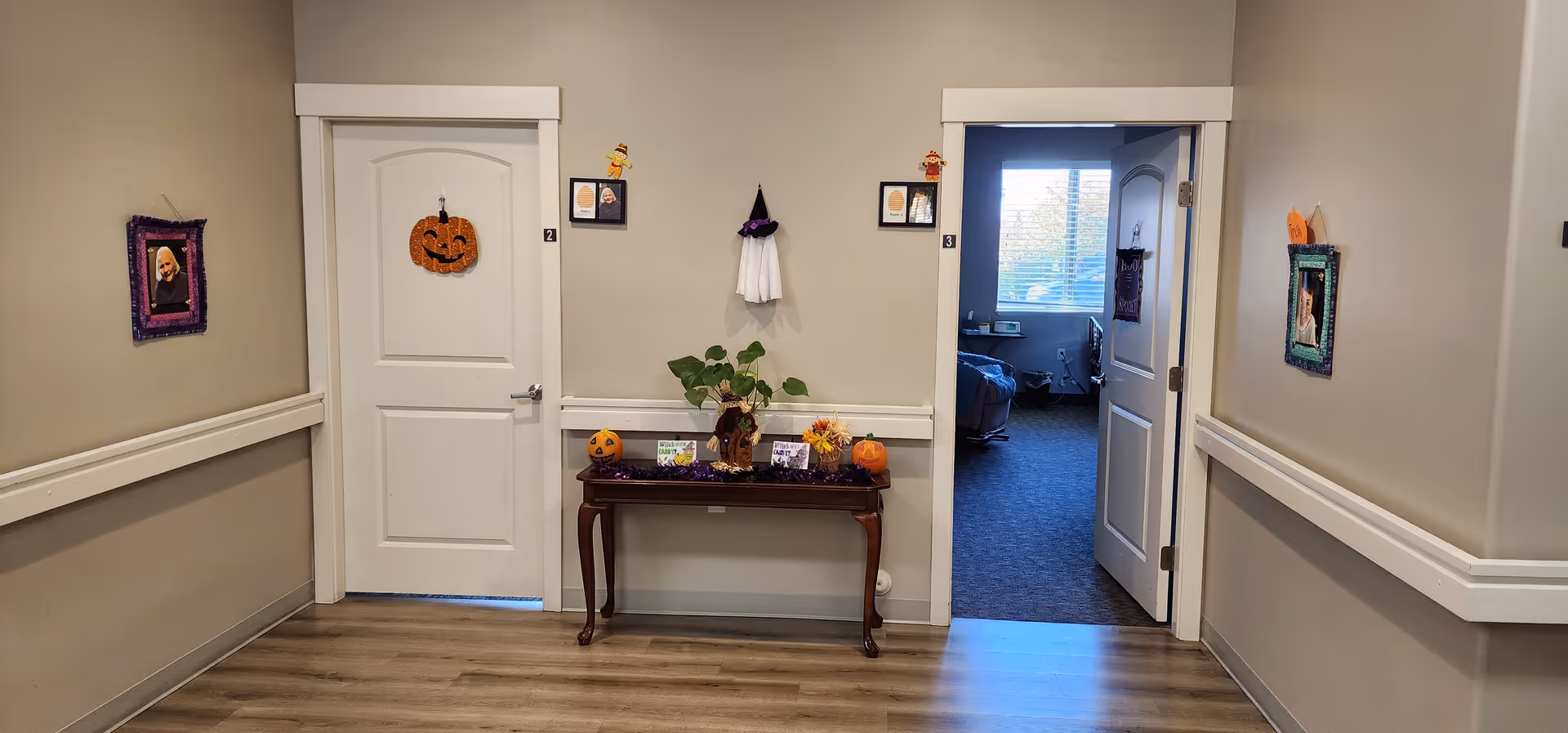 A hallway in a senior living facility with two white doors, one closed and one open showing a room with a bed and window. The hallway has light-colored walls and wood flooring. A small wooden table against the wall is decorated with Halloween-themed items including pumpkins and a small ghost figure. Framed photos and Halloween decorations hang on the walls around the doors.