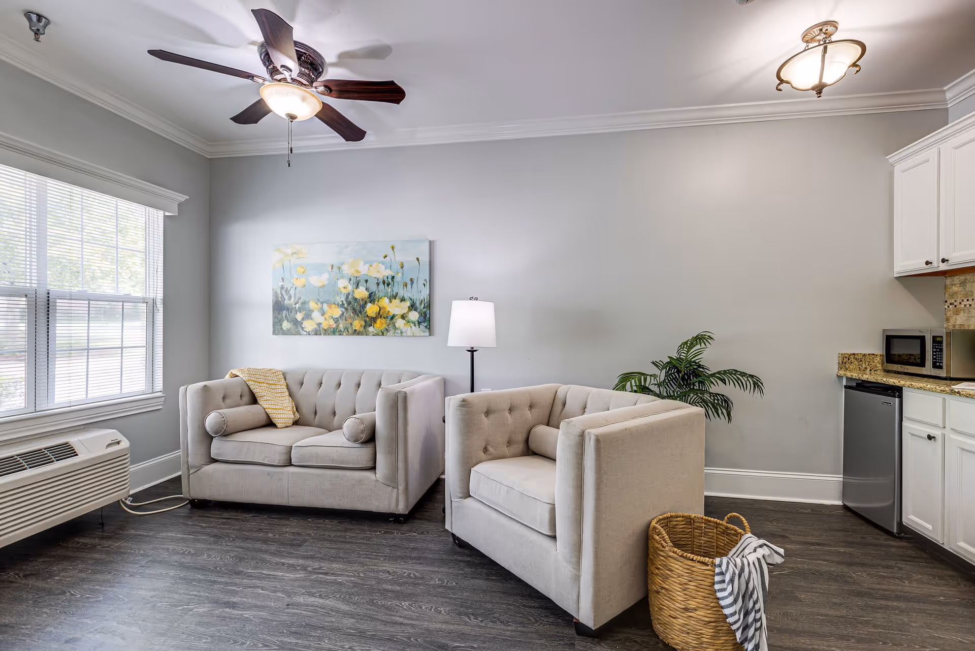 A cozy living room area with two beige tufted armchairs and a matching loveseat. A floor lamp stands between the chairs, and a large window with blinds lets in natural light. A ceiling fan with a light fixture is mounted on the ceiling. There is a framed floral painting on the wall above the loveseat. To the right, part of a kitchen area with white cabinets, a microwave, and a small refrigerator is visible. A woven basket with a striped cloth is on the dark wood floor.