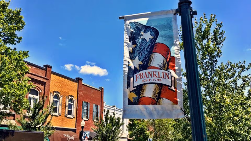 A street scene with a lamppost banner reading 'FRANKLIN EST. 1799' in front of brick storefronts, trees, and a blue sky.