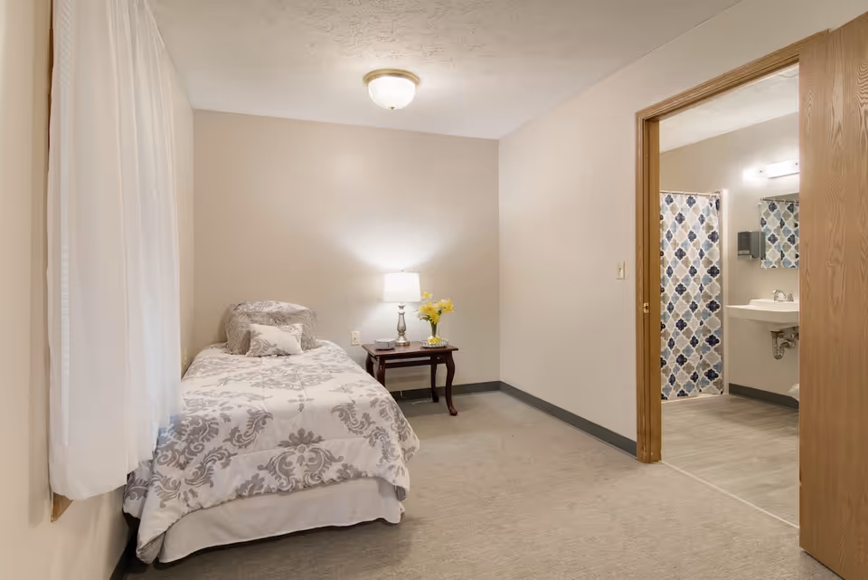 A small bedroom with a single bed covered in a white and gray patterned comforter. Next to the bed is a wooden nightstand with a lamp and a vase of yellow flowers. The room has beige walls and carpeted floor. An open door reveals a bathroom with a sink, mirror, and a shower with a blue and gray patterned curtain.