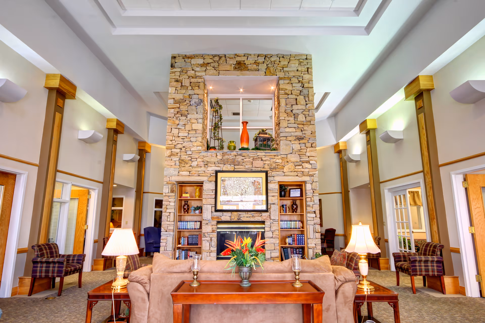 A spacious and well-lit living room area with a large stone fireplace as the focal point. The fireplace is flanked by built-in wooden bookshelves filled with books and decorative items. In front of the fireplace is a beige sofa with a wooden table behind it, holding a flower arrangement and two candle holders. There are two table lamps on side tables next to the sofa. The room has high ceilings with recessed lighting and several wooden chairs with plaid upholstery arranged along the walls. Multiple doorways lead to other rooms.