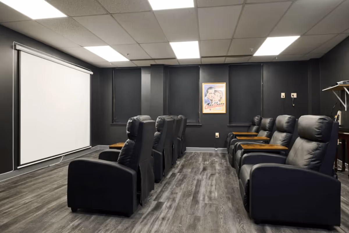 A small theater room with two rows of black leather recliner chairs facing a large white projection screen. The walls are painted black, and there is a framed vintage movie poster on the back wall. The floor has dark wood-style flooring, and the ceiling has white tiles with recessed lighting.