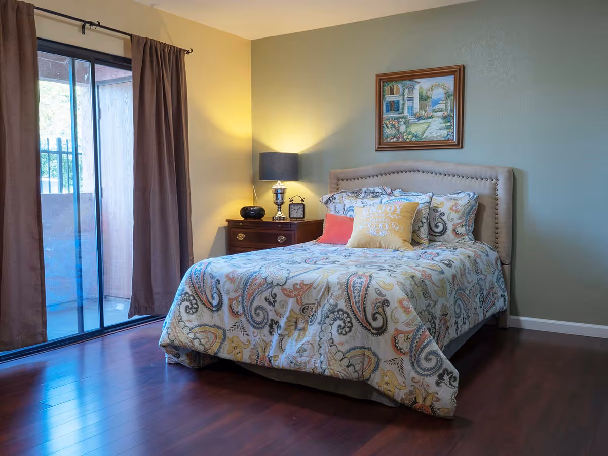 A neatly made bed with a paisley patterned comforter and multiple pillows in a bedroom. There is a wooden nightstand with a lamp, clock, and decorative item next to the bed. A framed painting hangs on the wall above the bed. The room has wooden flooring and a sliding glass door with brown curtains leading to an outdoor area.