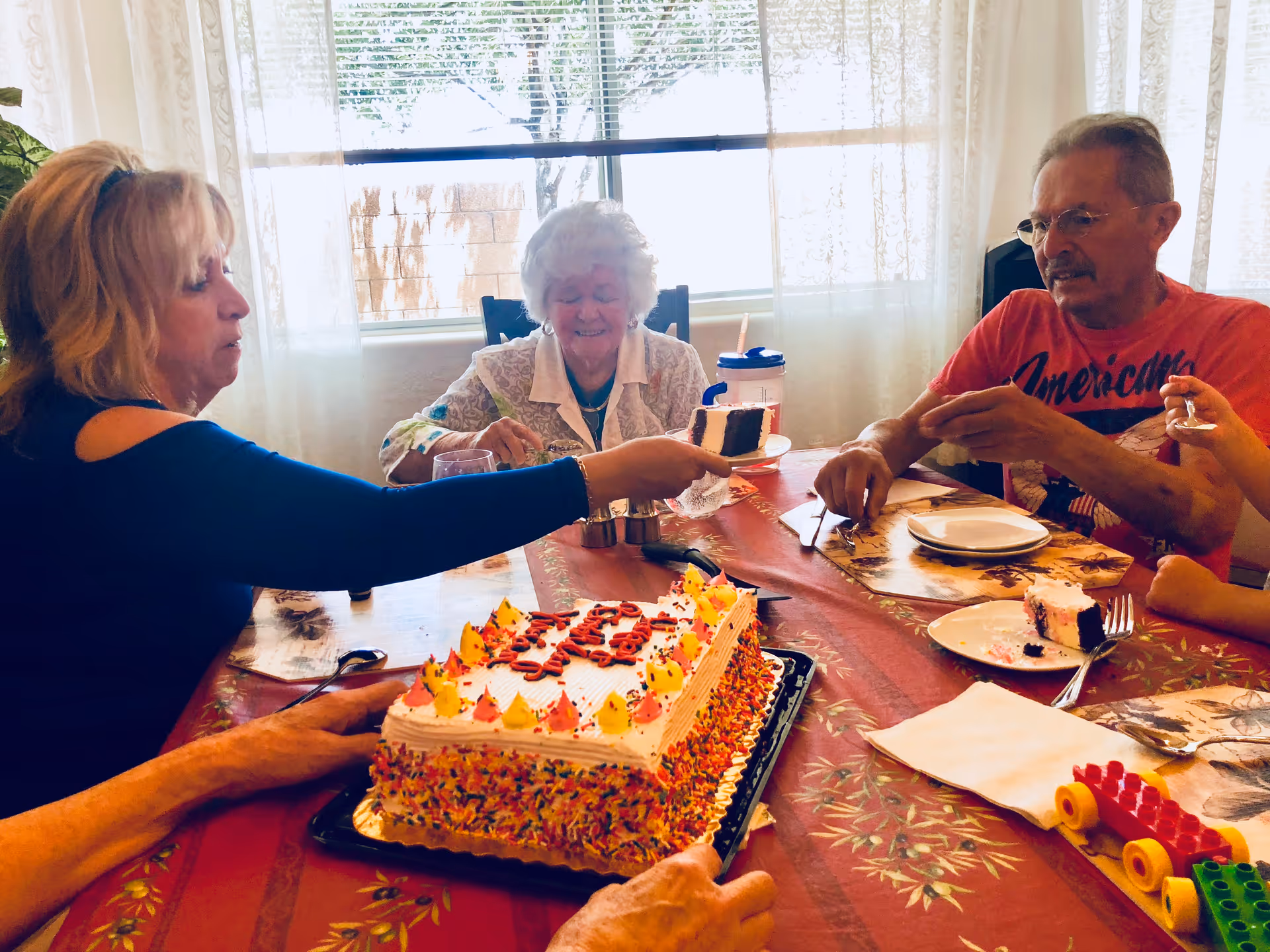 Several older adults sit around a table cutting and sharing a decorated sheet cake in a sunlit dining room.