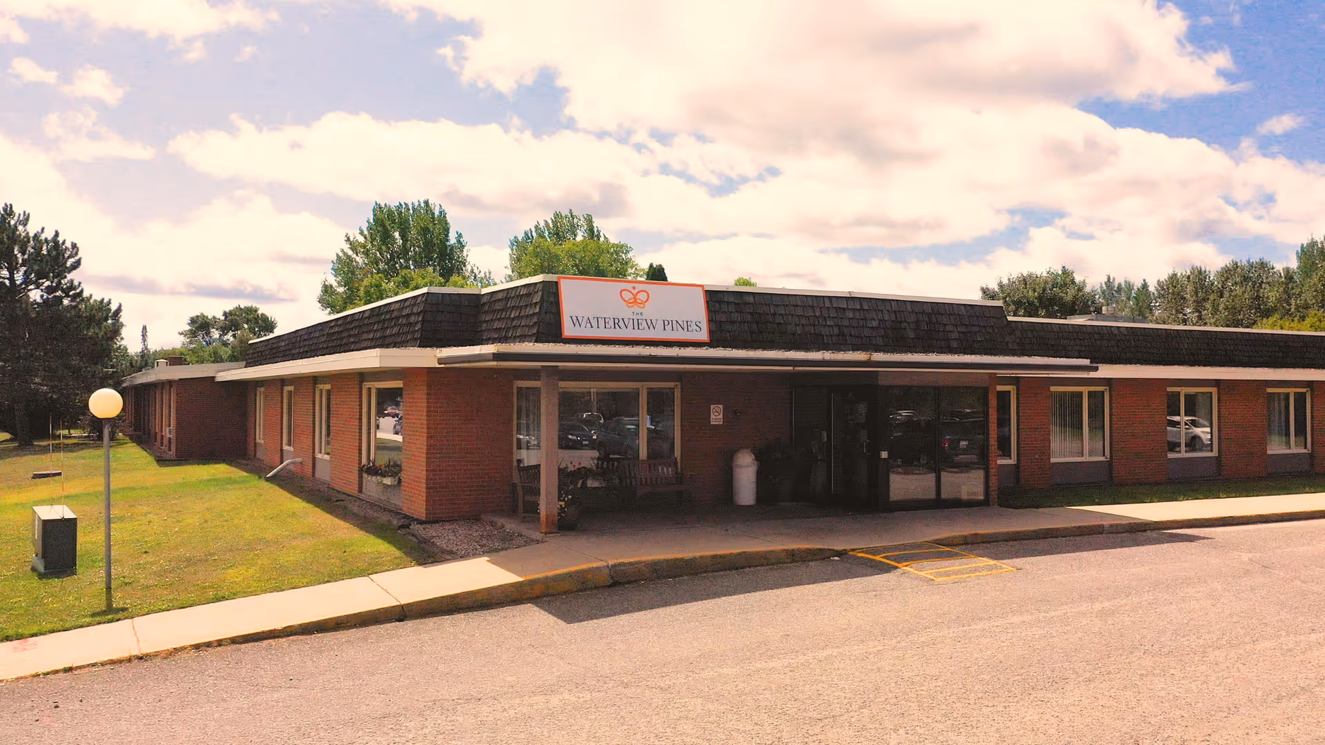 Exterior view of The Waterview Pines facility, a single-story brick building with a shingled roof, surrounded by green grass and trees under a partly cloudy sky. The entrance has a covered walkway and a sign above it displaying the facility's name.