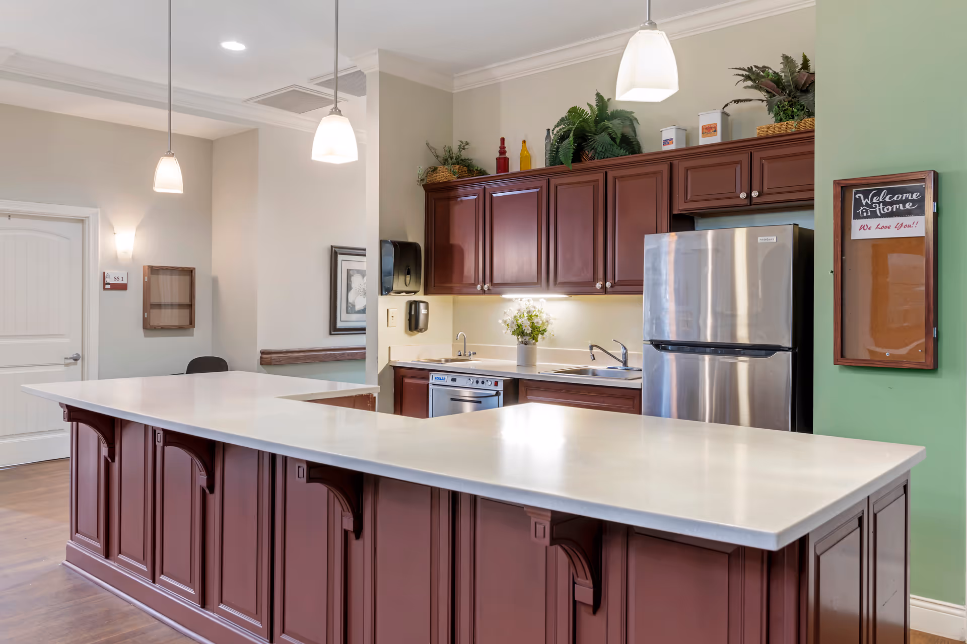 Interior view of a kitchen area in a senior living facility with a large white countertop island, dark wood cabinets, a stainless steel refrigerator, a sink, and under-cabinet lighting. There are three pendant lights hanging from the ceiling, plants and decorative bottles on top of the cabinets, and a small chalkboard on the green wall with the message 'Welcome Home We Love You!!'.