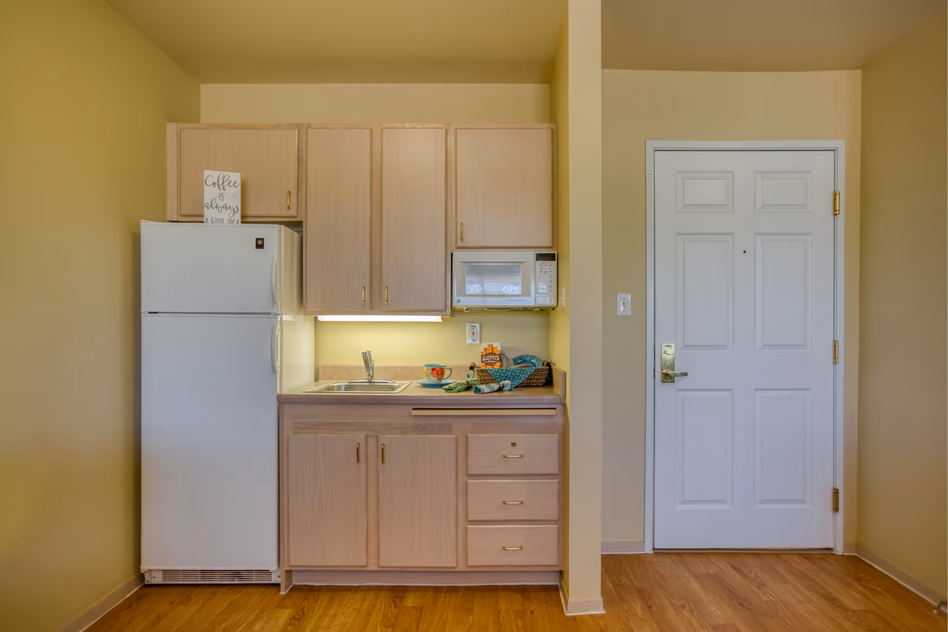 A small kitchen area with light wood cabinets, a white refrigerator, a microwave, and a sink. There is a sign on top of the refrigerator that says 'Coffee is always a good idea.' To the right of the kitchen is a white door with a gold handle. The floor is wooden and the walls are painted a light yellow color.