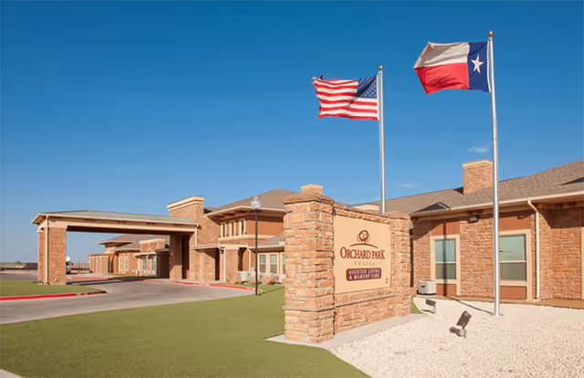 Exterior view of Orchard Park of Permian Basin assisted living and memory care facility with a stone sign in front, two flagpoles displaying the American and Texas flags, and a covered entrance to the building under a clear blue sky.