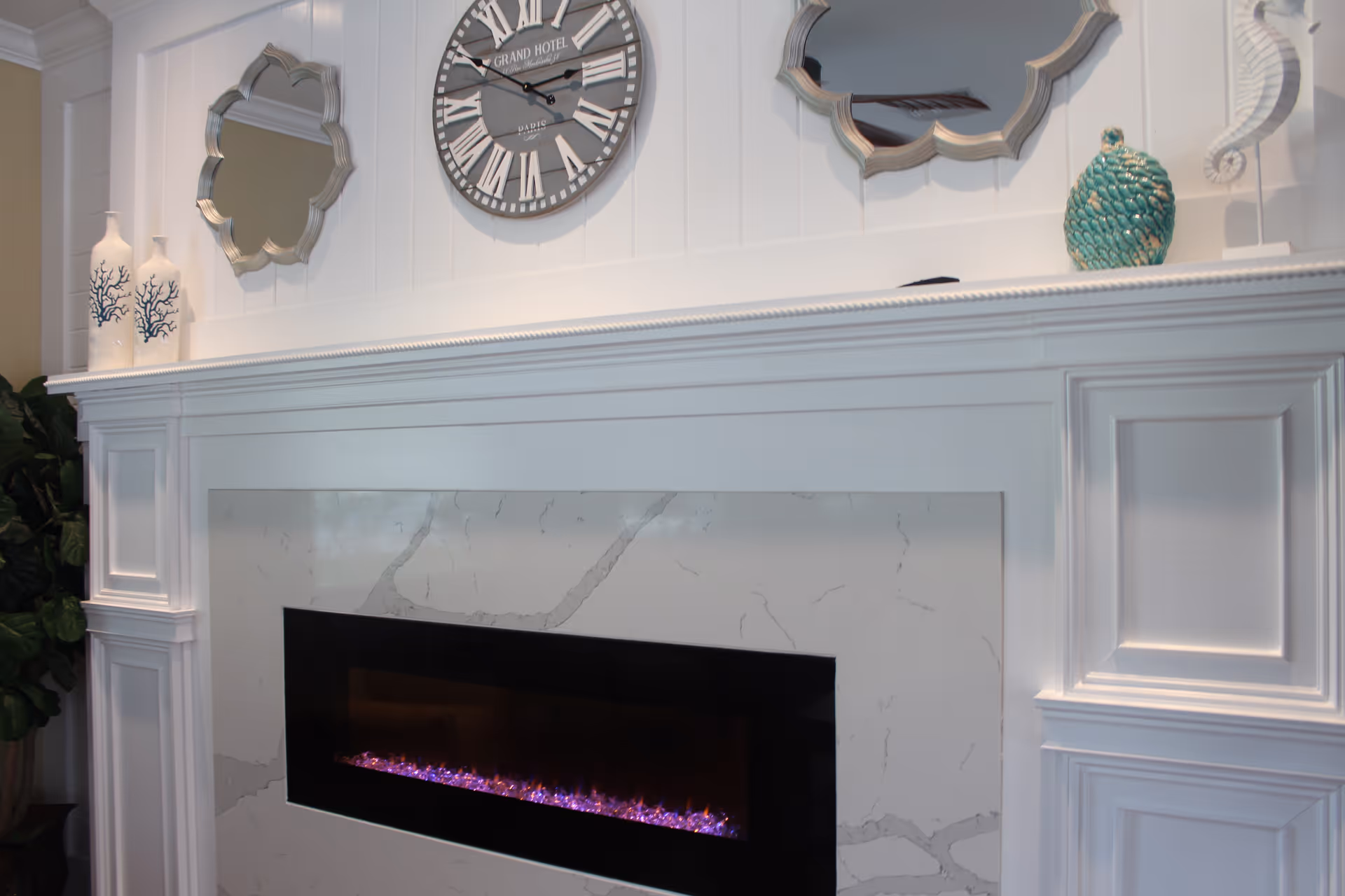 A white fireplace with a modern electric fire insert featuring purple flames. Above the fireplace are decorative items including two white vases with blue coral designs, a large round wall clock with Roman numerals, two decorative mirrors with scalloped edges, a turquoise textured vase, and a white seahorse figurine. There is also a green leafy plant partially visible on the left side.