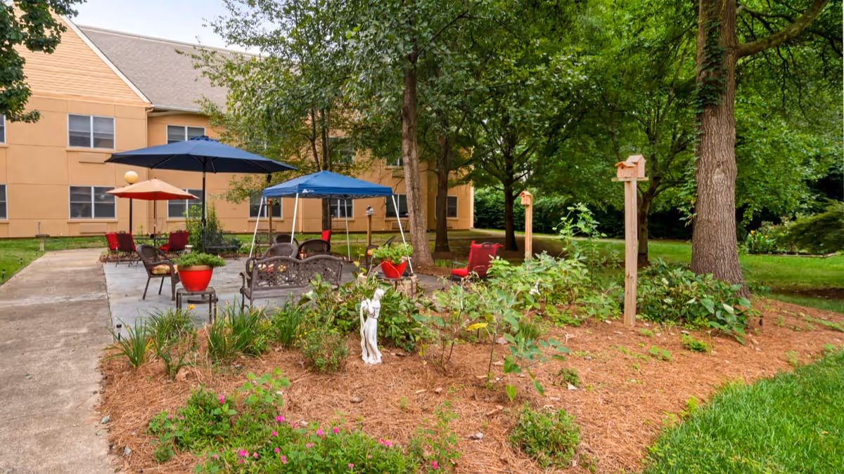 Outdoor patio area at Brookdale Charlotte East with several seating arrangements including chairs and tables under blue and red umbrellas. The patio is surrounded by trees, plants, and a garden bed with a small white statue and birdhouses on wooden posts. The building exterior is visible in the background.