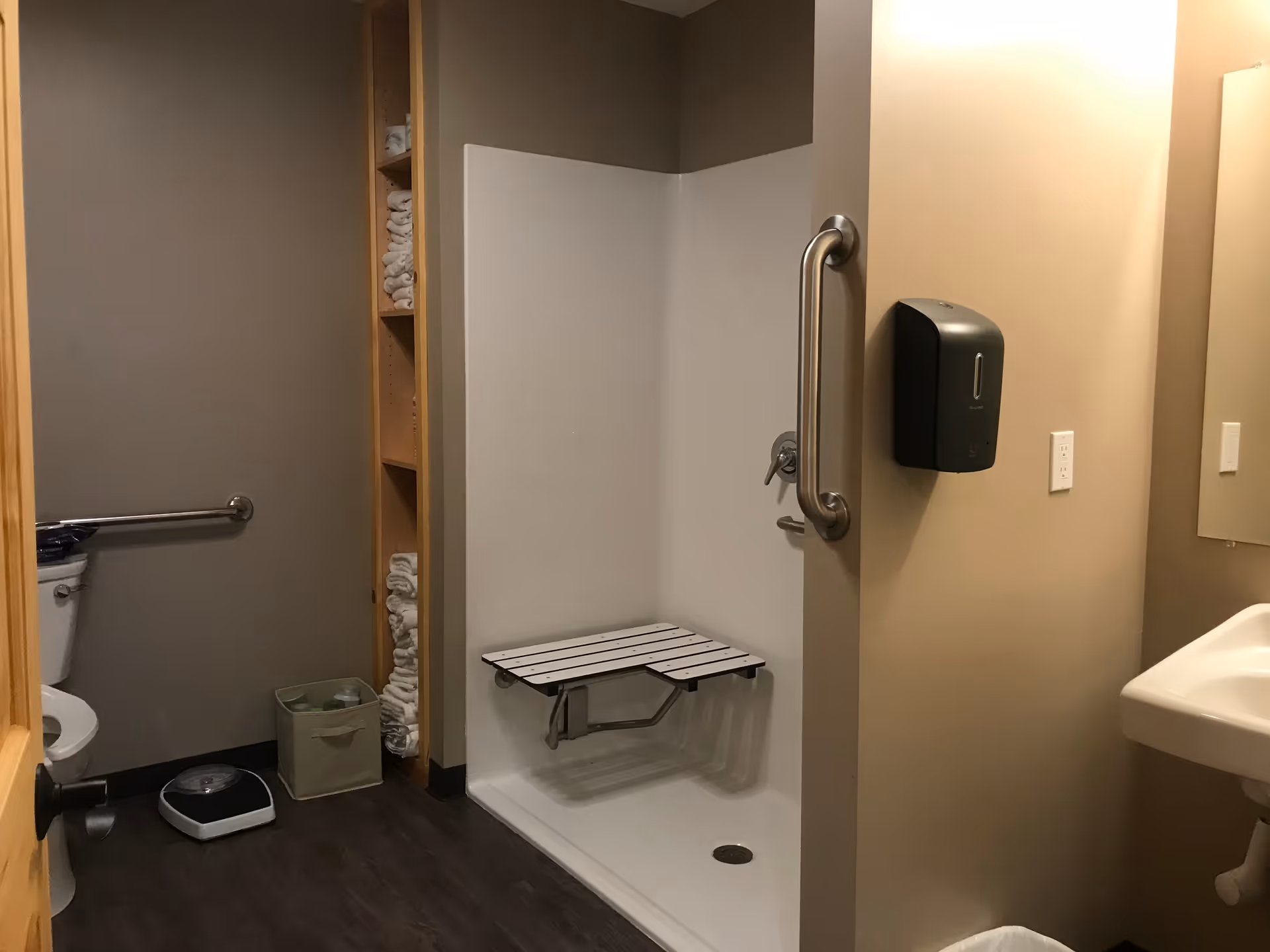 Accessible bathroom with a toilet, a wall-mounted grab bar, a shower area with a foldable seat and grab bar, a sink with a soap dispenser mounted on the wall, and shelves stocked with folded towels.