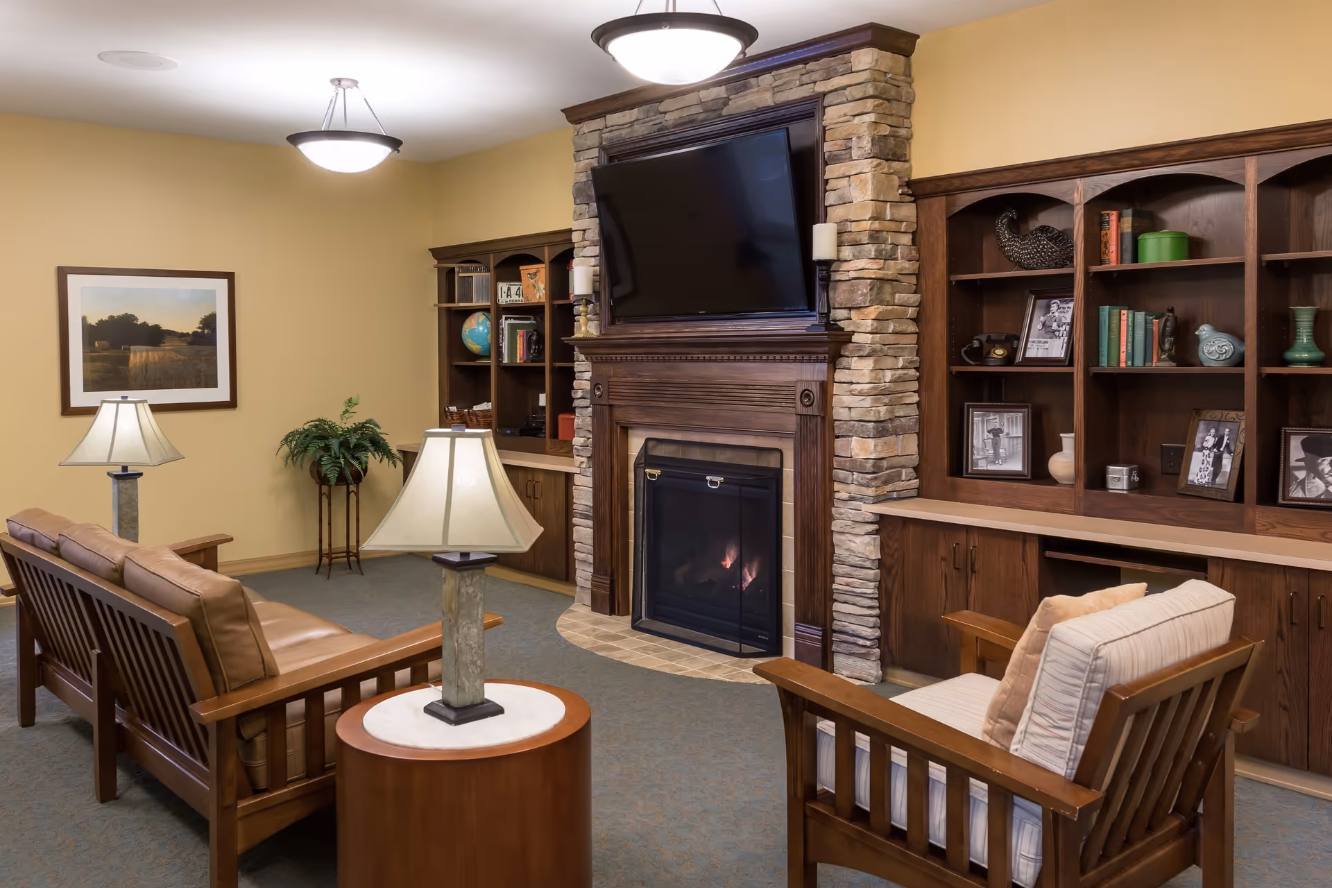 A cozy living room with a stone fireplace and a mounted flat-screen TV above it. The room features wooden bookshelves filled with books, framed photos, and decorative items. There are two wooden armchairs with cushions and a wooden sofa with leather cushions arranged around a small round table with a lamp. The walls are painted beige, and the room is softly lit by ceiling lights and table lamps.