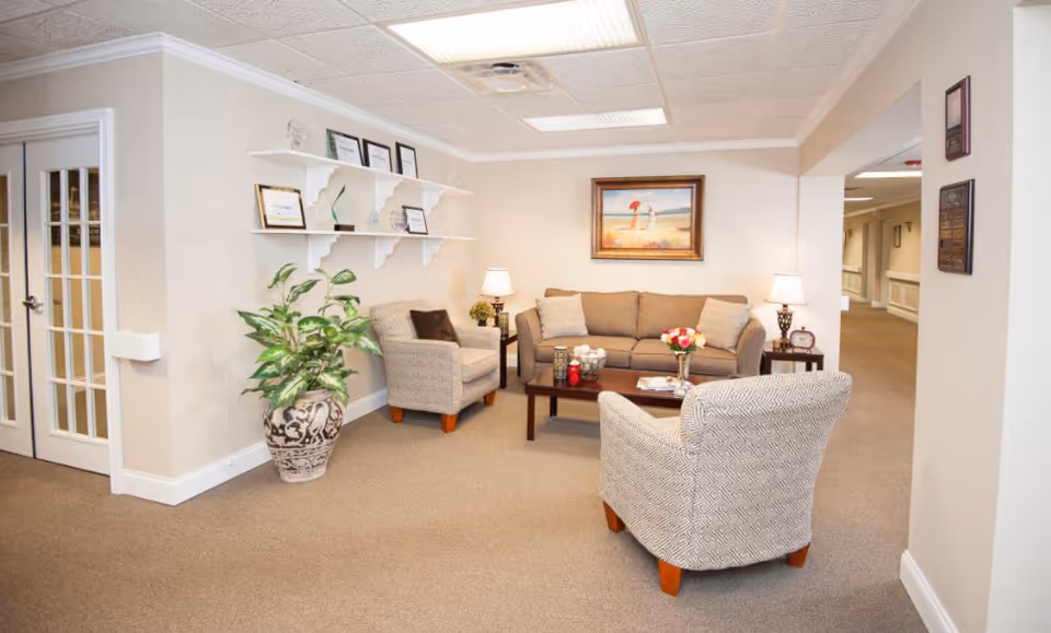 A cozy seating area in a senior living facility with a beige sofa, two patterned armchairs, a wooden coffee table with decorative items, two table lamps, a large potted plant, and framed certificates on white shelves mounted on the wall. A framed painting of a beach scene hangs above the sofa. The room has beige walls and carpet, with a hallway visible in the background.