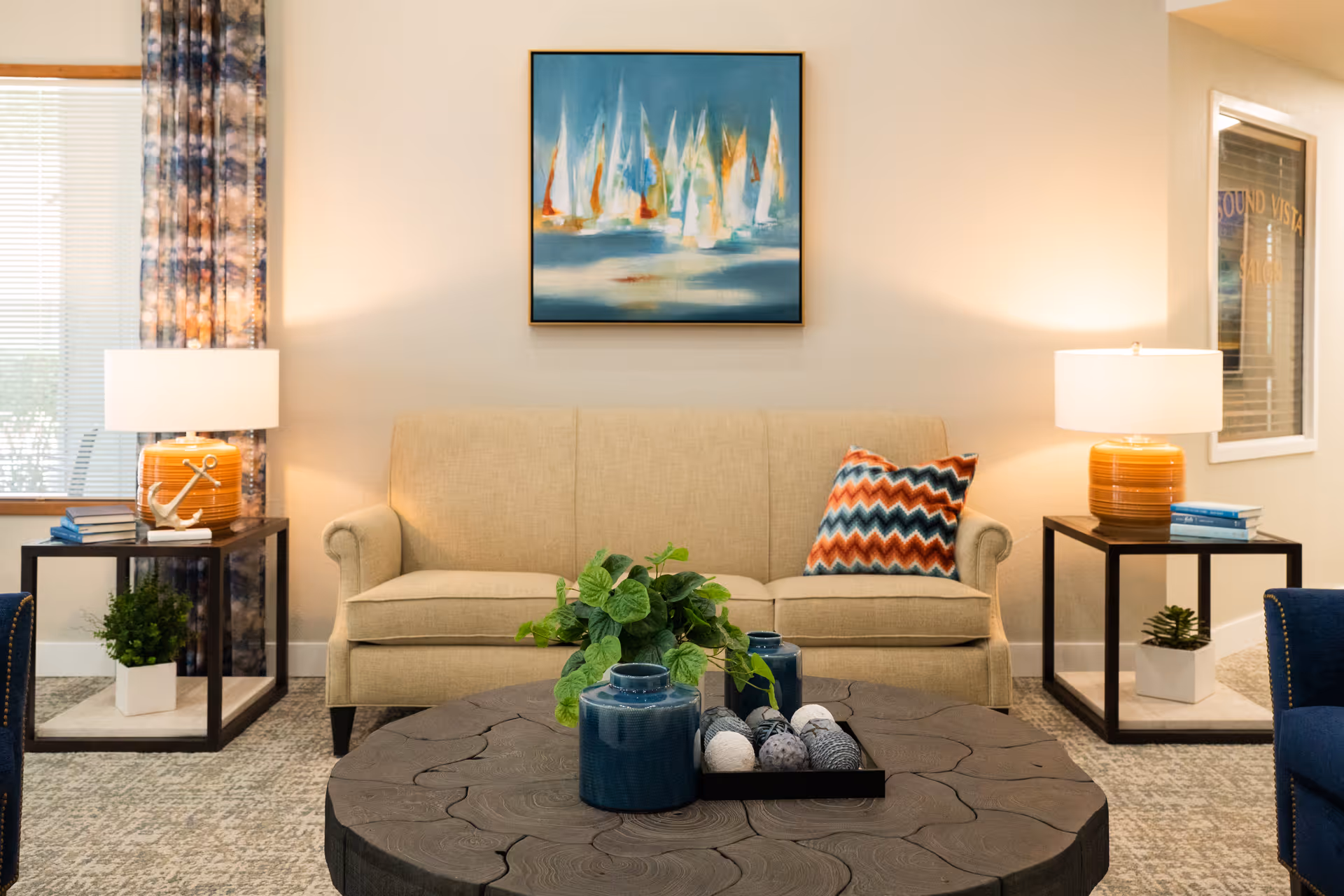 A cozy living room with a beige sofa adorned with a colorful zigzag patterned pillow. On either side of the sofa are black side tables with orange lamps and small potted plants. A round wooden coffee table with decorative vases and a plant is in the foreground. A painting of sailboats hangs on the wall above the sofa, and a window with patterned curtains is visible to the left.